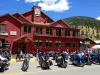 A large, two-story red building housing Texas Reds Steakhouse and The Lodge at Red River, featuring a row of motorcycles parked along the street front. People are walking on the sidewalk and dining on the patio under a bright blue sky with forested mountains in the background.