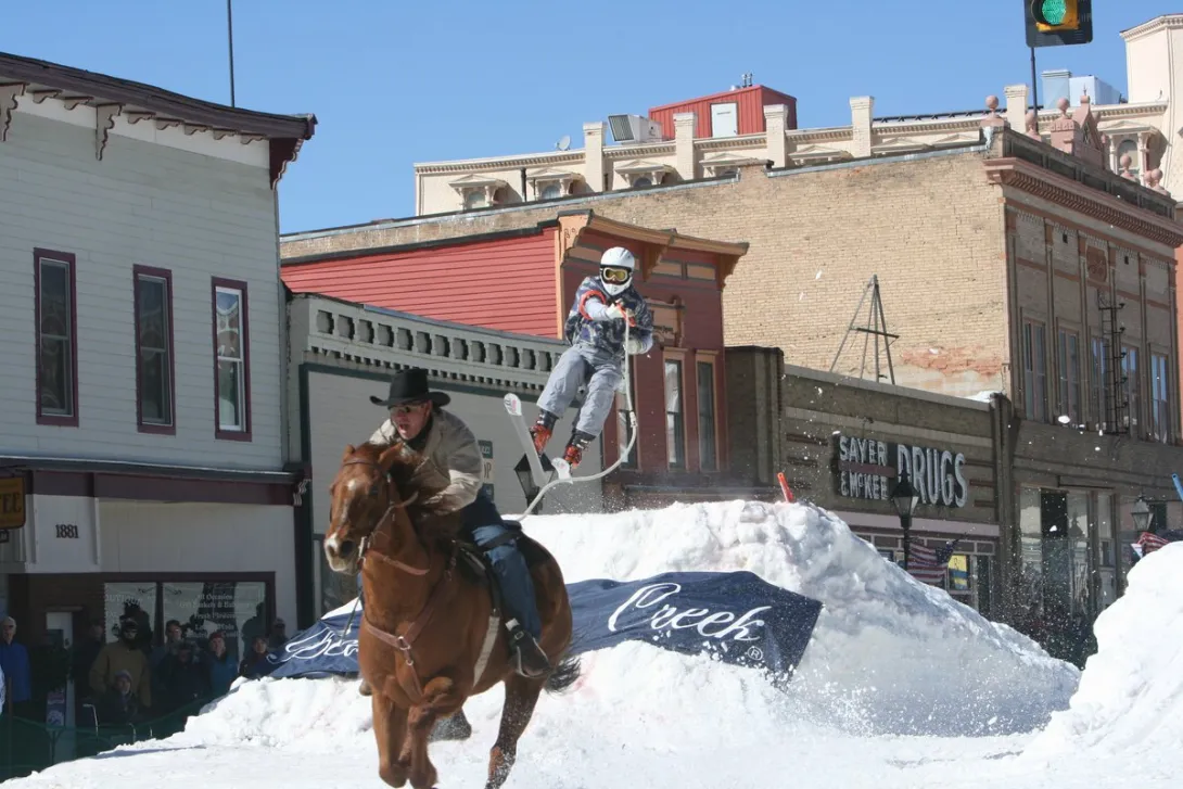 A horse at full gallop pulls a skier over a snow jump during a skijoring race.