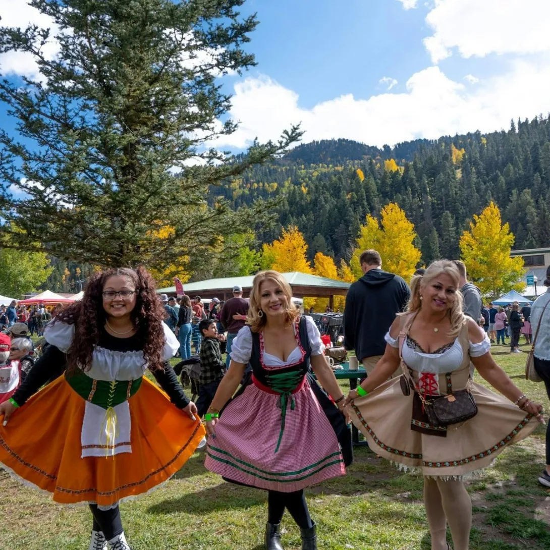 Women in dirndls celebrate at Red River Oktoberfest in Brandenburg Park beneath golden aspens.