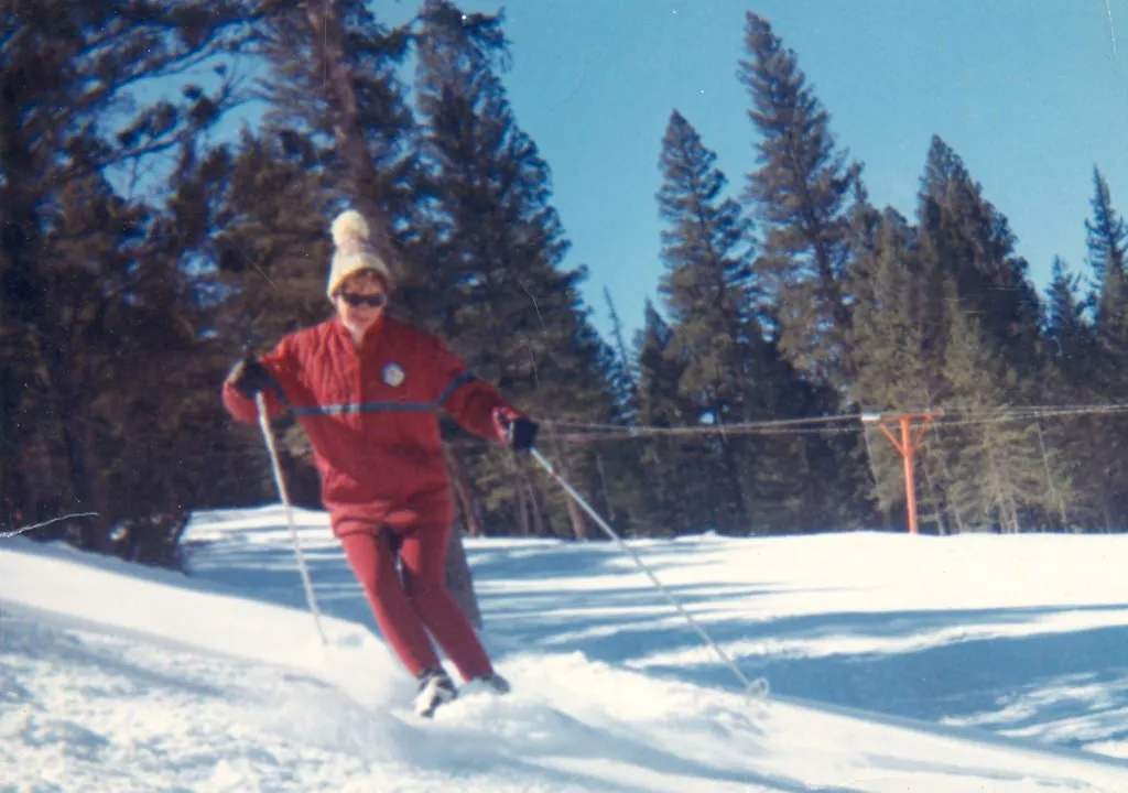 Judy Miller skiing at Powder Puff Mountain in Red River in the 1970s, in a red ski suit with a rope tow behind her.