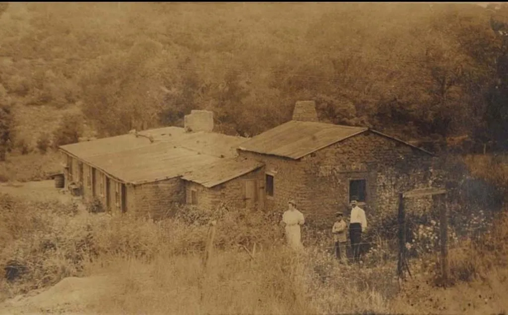 A sepia-toned historic photograph of Richens L. Wootton's stone house at Raton Pass, with three people standing in front.